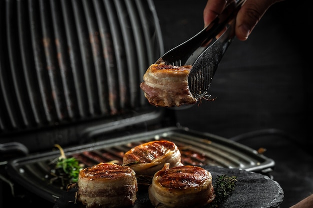 Photo medallions steaks from the beef tenderloin covered bacon on grill with smoke dark background. cooking beef steak on grill by chef hands.