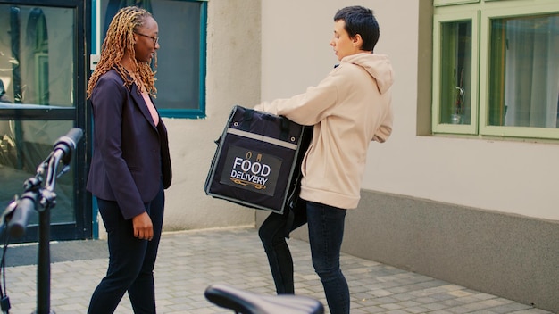 Medewerker afhaalmaaltijden bezorgt fastfoodbestelling aan klant die bij de ingang wacht en geeft restaurantpakket. Vrouwelijke bezorger met papieren zak met lunchmaaltijd, vrouw op fiets.