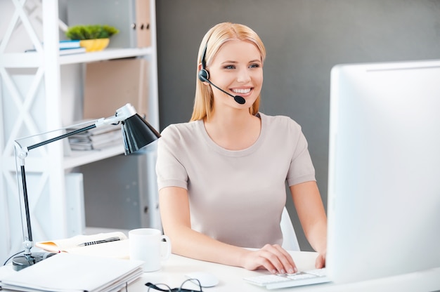 Medewerker klantenservice op het werk. Mooie jonge vrouw in hoofdtelefoon die op de computer werkt en glimlacht terwijl ze op haar werkplek op kantoor zit