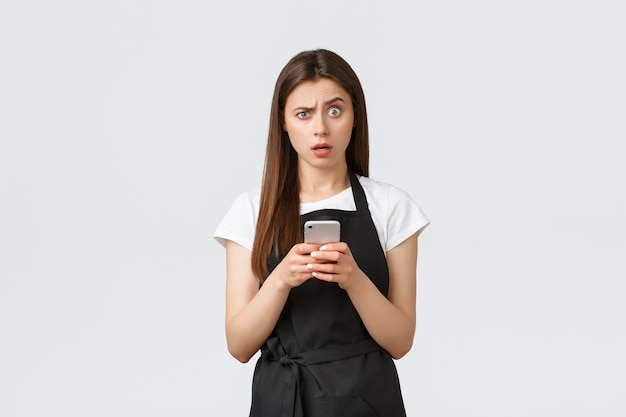 Medewerkers van supermarkten, kleine bedrijven en coffeeshops concept. Verward en besluiteloos vrouwelijke barista in zwarte schort wenkbrauw twijfelachtig, met behulp van mobiele telefoon, vreemd bericht ontvangen.