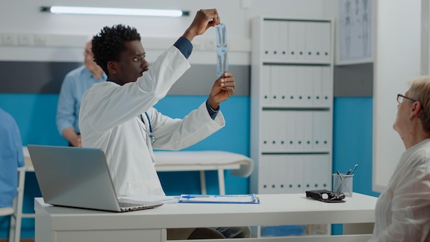 Medic of african american ethnicity analyzing x ray of patient healthcare sitting at desk. Black doctor holding radiography scan for professional diagnosis and results in medical cabinet