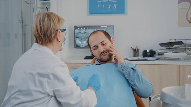 Фото Медик консультирует пациента с проблемами зубных протезов и зубной болью, сидящим на стоматологическом кресле. стоматолог проводит обследование для лечения человека с кариесом, говорит о хирургической процедуре.