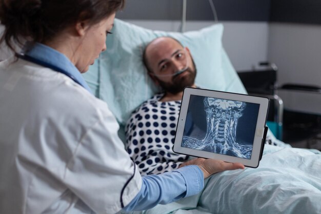 Медик объясняет травму шейки матки мужчине средних лет с низкой сатурацией spo2 на больничной койке. Врач смотрит на цифровой планшет с МРТ диагностики чтения горла пациенту, получающему кислород.