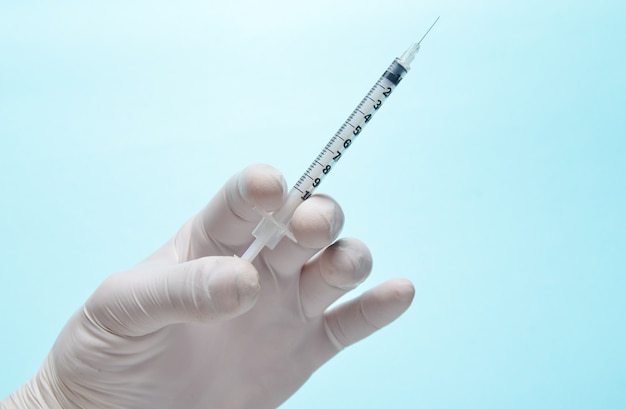 Medic's hand with white latex gloves holds syringe with needle on blue.