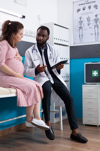 Medic showing digital device with diagnosis to pregnant patient, checking healthcare in office. General practitioner using tablet to examine young pregnant woman. Medical care