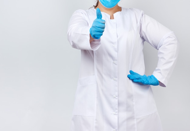 Photo medic woman in white coat and mask, wearing blue medical latex gloves on her hands, showing like gesture in hand