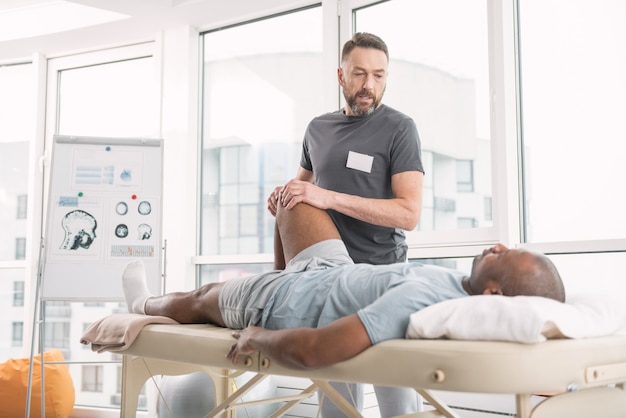 Medical aid. Smart nice skilled doctor looking at his patient while holding his leg