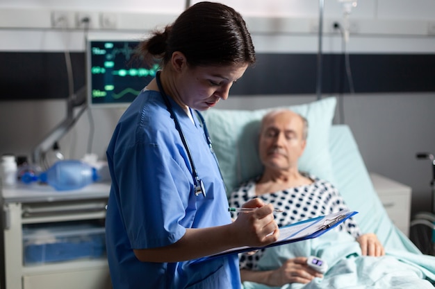 Medical assistant checking treatment of senior man reading notes on clipboard sick elderly laying in...