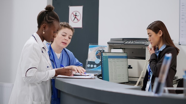 Medical assistant and doctor analyzing report papers at reception counter, working on checkup forms and appointments to help patients. Diverse staff at hospital registration desk in lobby.