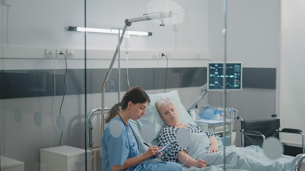 Medical assistant doing checkup visit for patient in hospital\
ward bed. woman working as nurse consulting aged woman with iv drip\
bag and nasal oxygen tube to cure disease with treatment