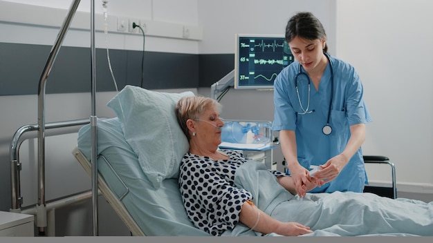 Medical assistant putting oximeter on finger of aged patient in\
hospital ward bed for oxygen saturation measurement and pulse\
pressure. nurse helping retired woman with iv drip bag