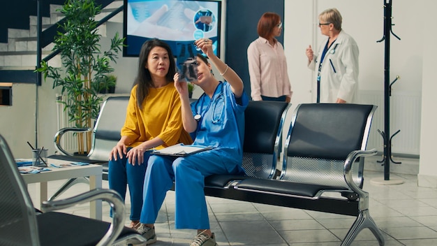 Medical assistant showing x ray scan to elderly asian woman in\
waiting area lobby, attending checkup examination at clinic. nurse\
and patient looking at radiography results diagnosis.