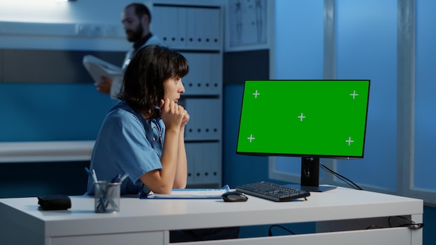 Medical assistant standing at desk looking at computer with\
green screen display working late at night during checkup visit\
appointment. physician nurse typing patient expertise in hospital\
office