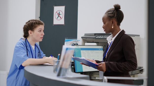 Medical assistant talking to receptionist about appointments\
and consultation at reception desk. diverse hospital staff working\
on report papers, filling in medical forms at registration\
counter.