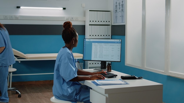 キャビネット内のコンピューターのキーボードで入力する医療助手。机に座ったまま、健康診断や医療行為のためにモニターを使用して制服を着た看護師として働く女性。