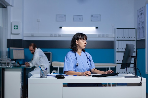 의료 시스템을 위해 사무실에서 키보드와 컴퓨터를 사용하는 의료 보조. 여성 간호사는 도움과 지원을 위해 모니터 화면을 보고 밤 늦게까지 일합니다. 건강 전문가
