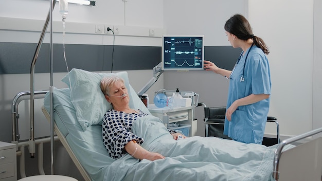 Medical assistant visiting retired patient sleeping in hospital ward bed. Woman working as nurse checking on heart rate monitor for pulse measurement. Specialist taking care of person