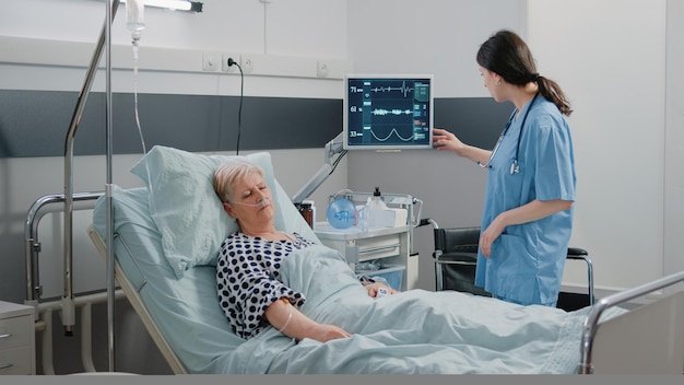 Medical assistant visiting retired patient sleeping in hospital\
ward bed. woman working as nurse checking on heart rate monitor for\
pulse measurement. specialist taking care of person