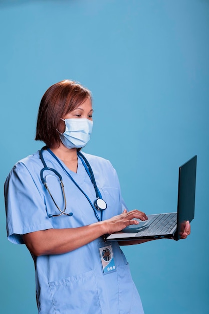 medical assistant wearing protective face mask to prevent infection with covid19 holding laptop computer checking virus expertise. nurse in blue uniform working at diagnosis during checkup visit