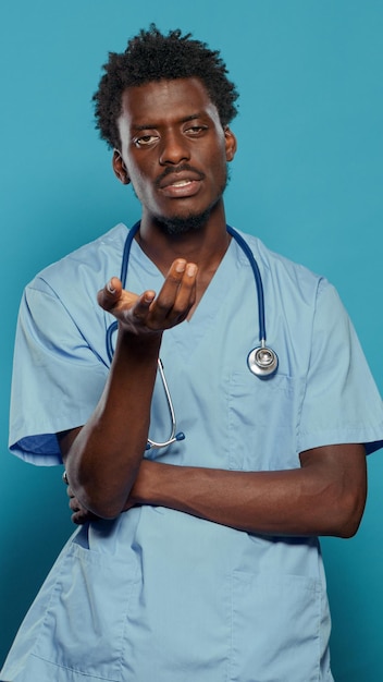 Medical assistant wearing uniform and stethoscope while talking and looking at camera in studio. Man nurse explaining healthcare while standing over isolated background. Health specialist