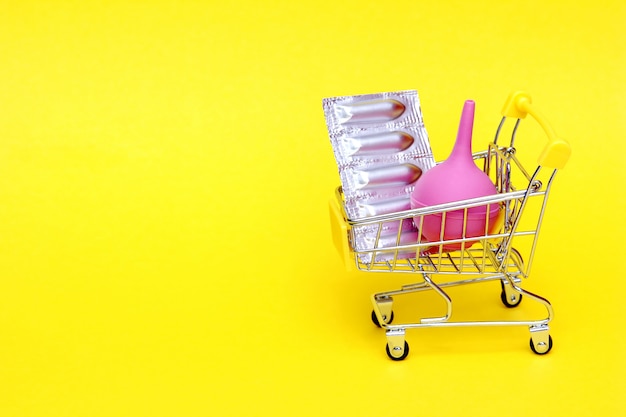 Medical candles in a silver blister pack and a pink rubber medical enema are placed in a toy shopping cart. Packaging of contraceptives, vaginal, rectal candles. Enema in medicine.