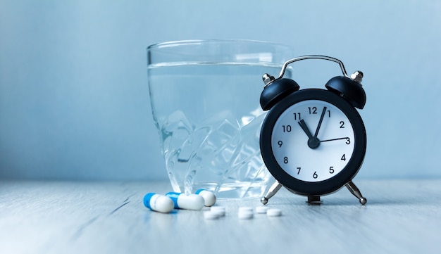 Medical capsule pills, glass water and alarm clock on blue background. Concept health 