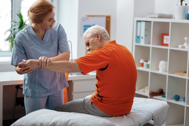 Medical center. Delighted friendly nurse standing in front of her patient while holding his hand