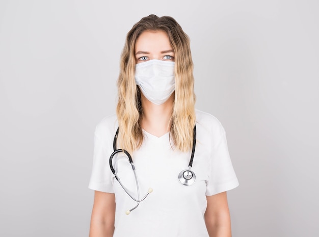 Medical concept of beautiful woman doctor in white coat with stethoscope, mask. Medical student. Woman hospital worker and smiling, gray wall