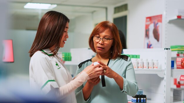 Medical consultant showing cardiology pills to asian woman in\
pharmacy, explaining treatment to cure heart problems. female\
pharmacist talking to client about medical products. handheld\
shot.