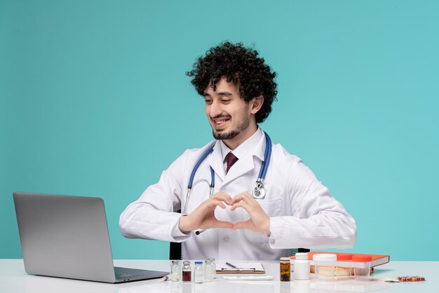 Medical cute handsome doctor working on computer remotely in lab coat showing heart gesture