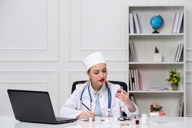 Medico adorabile sveglio medico in camice bianco e cappello con il computer che esamina le pillole