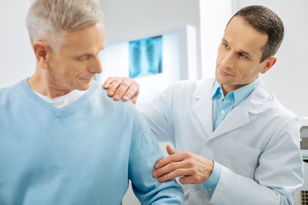 Medical diagnostics. Professional nice mart doctor standing near his patient and checking his arm while working with him