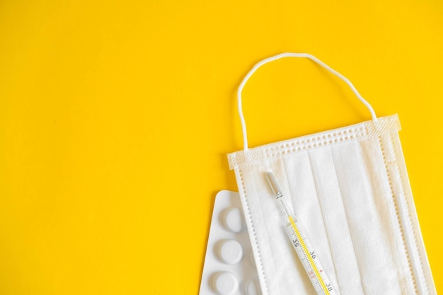 Medical disposable mask, pills and a mercury thermometer on a yellow background. Top view. Copy, empty space for text