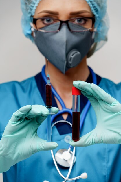 Medical doctor nurse woman wearing protective mask and gloves - holding virus blood test tube and syringe