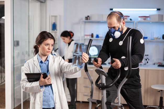 Medical doctors in sports science lab measuring performance cardiorespiratory of athlete. Team of researcher monitoring vo2 of sportsman running on cross trainer equipped with electrodes and mask.