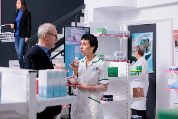 Foto impiegato medico che mostra pillole per il cuore del cliente senior e fornisce raccomandazioni nel negozio al dettaglio della farmacia. lavoratore farmaceutico e uomo anziano in piedi vicino allo scaffale dei farmaci, discutendo di assistenza sanitaria