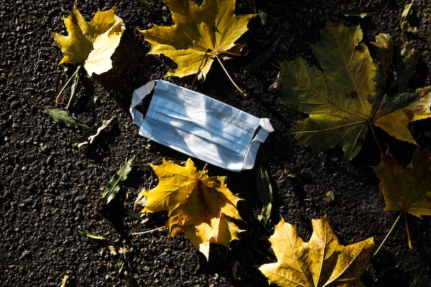 Medical face mask lying on asphalt with autumn leaves outdoors