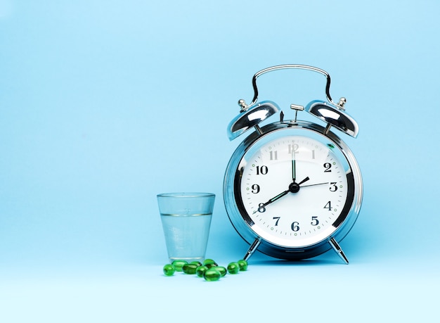 Medical green capsule pills, glass water and alarm clock on blue background. Concept health