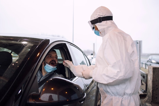 Медицинский работник в белом защитном костюме, перчатках и маске берет мазок из носа и горла для проверки пассажира на вирус короны.