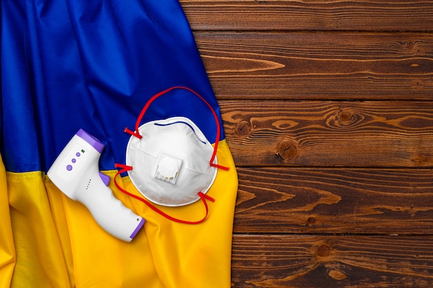 Medical mask and contactless thermometer on the flag of Ukraine on wooden board