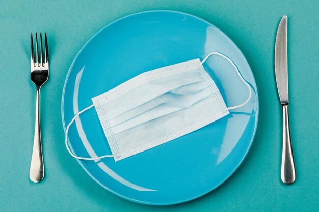 A medical mask on an empty plate with a fork and knife on a blue background The concept of nutrition and protection against the virus