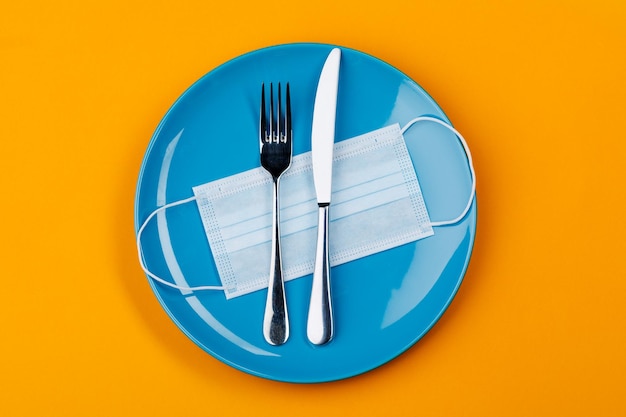 A medical mask on an empty plate with a fork and knife on an orange background