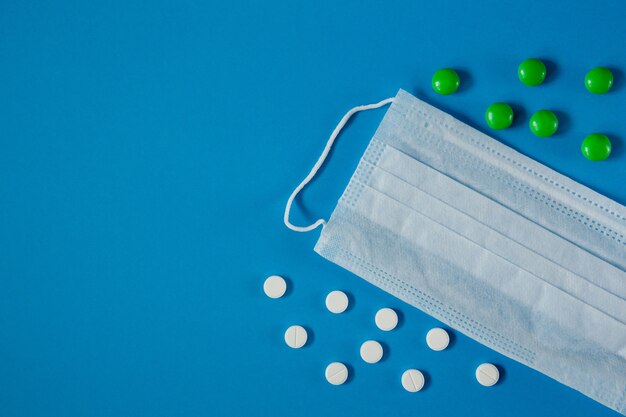 Medical mask on face and pills on blue table from above