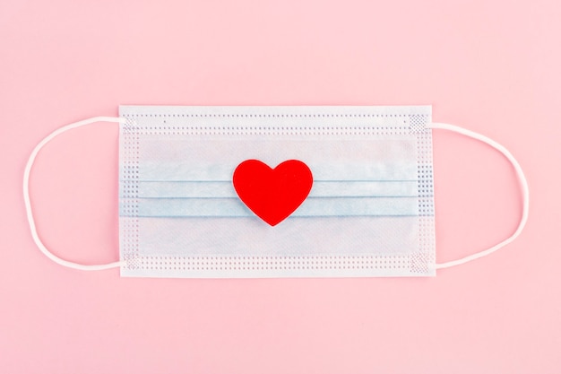 medical mask on pink background, red heart, top view