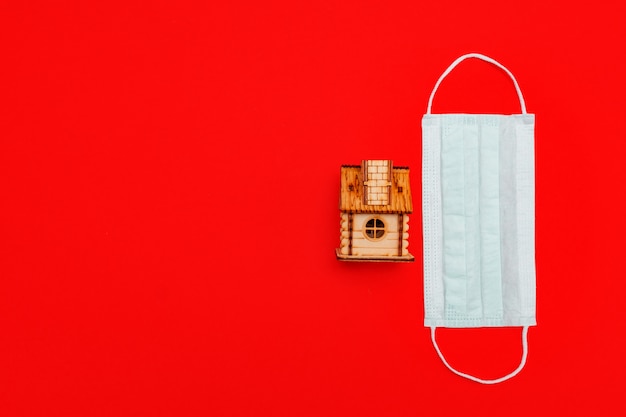 Medical mask and wooden toy house on red backgound. concept of self quarantine at home as preventative measure against virus outbreak.