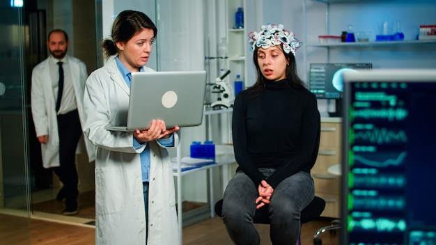 Photo medical neurologist doctor asking symptoms of patient taking notes on laptop, woman wearing high tech brain scanner