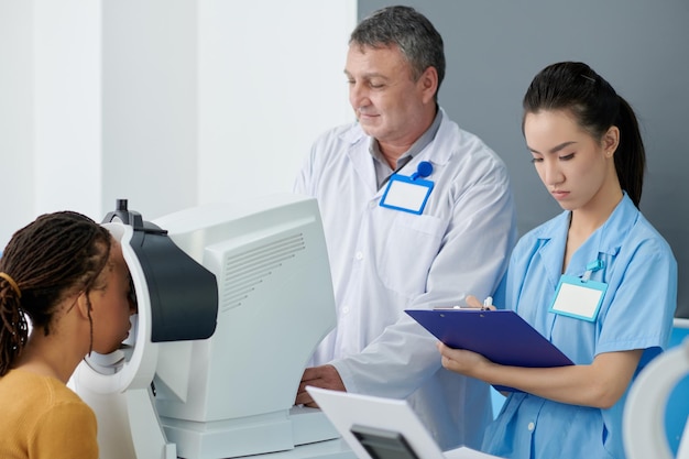 Medical nurse taking notes in document when doctor examining eyes of patient with moden equipment