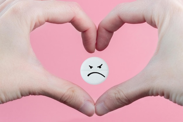 Medical pill with a sad emoticon in the heart from the hands on a pink background Health care