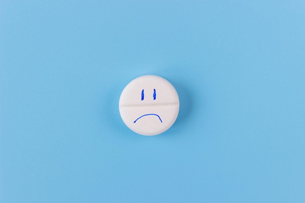 Медицинская таблетка с грустным лицом на синем фоне. Концепция здравоохранения или медицинской помощи.