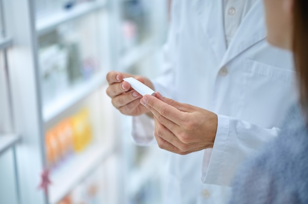 Medical product. Pharmacist consultant hands in white coat holding small tube showing visitor in pharmacy, face is not visible